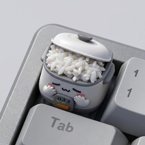 Puede incluir: Una tecla personalizada con forma de olla arrocera, llena de arroz blanco, sobre un teclado de ordenador. La tecla es blanca y gris con un diseño de dibujos animados. La tecla está en la tecla Tab.