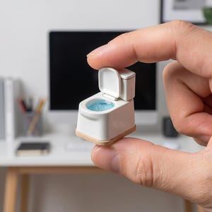 May include: A miniature white toilet with the lid open, revealing a blue interior. The small toilet has a light brown base and is held between fingers. The background includes a computer monitor and a desk.