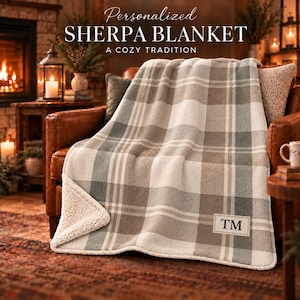 May include: A personalized sherpa blanket featuring a grey, white, and beige plaid pattern with a white fleece underside. A rectangular patch on the corner displays the letters TM in black text. The blanket is draped over a brown leather chair.