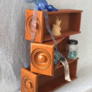 May include: A three-tiered wooden shelf unit with a warm brown finish. Each shelf features a decorative circular design. The shelves are adorned with a blue glass ornament, a small bear-shaped candle, and a glass jar.