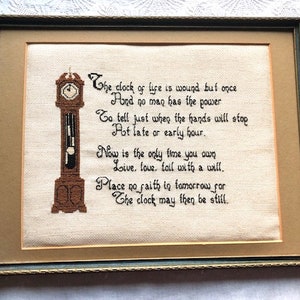 May include: Framed cross-stitch artwork featuring a brown grandfather clock design and a poem in black script. The artwork is framed in a gold-toned mat and a dark green and gold frame. The text reads "The clock of life is wound but once..."