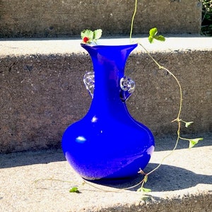 May include: A cobalt blue glass vase with clear glass handles. The vase has a rounded body and a narrow neck. It is sitting on a concrete step with a green vine growing up the side.
