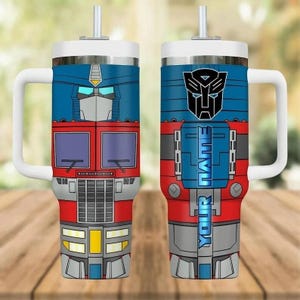 May include: A double-walled tumbler featuring a design inspired by Optimus Prime from Transformers. The tumbler has a white handle and lid, a blue and red color scheme, and the text "YOUR NAME" in a vertical format. The design includes the Autobot symbol.