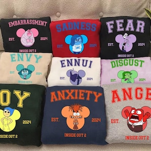 May include: A collection of Inside Out 2 themed sweatshirts in various colors, each featuring a different emotion: Embarrassment, Sadness, Fear, Envy, Ennui, Disgust, Joy, Anxiety, and Anger. Each sweatshirt has the emotion's name, a character graphic, and the text "EST. 2024 INSIDE OUT 2".