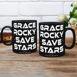 May include: Two black coffee mugs with white text that reads "GRACE ROCKY SAVE STARS" and "PROJECT HAIL MARY". The mugs are on a wooden surface with a white brick background.