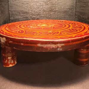 May include: A round, low wooden table with a red and orange decorative design on the top. The table has three short legs and a distressed finish, giving it a vintage appearance. The design features geometric and floral patterns.