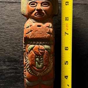 Puede incluir: Estatua de color terracota de una figura con tocado, de aproximadamente 28 cm de altura. La estatua tiene tallas detalladas y detalles verdes. La figura tiene una base rectangular y está de pie.