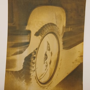 May include: Sepia-toned photograph of a car's tyre and wheel, with a portion of the car's body visible. The tyre has a textured tread pattern, and the wheel has a circular design. The image is vintage, with a sepia colour scheme.