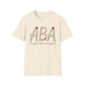 以下が含まれることがあります： 赤と緑の花柄のデザインで「ABA」の文字がプリントされたクリーム色のTシャツ。「applied behavior analysis」の文字が筆記体で書かれています。