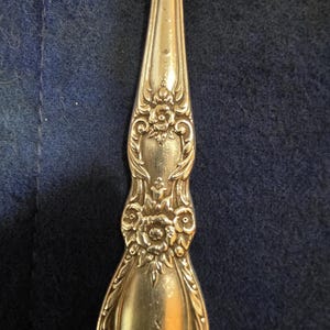 Peut inclure: Gros plan d'une cuillère dorée avec des détails floraux et des volutes ornés. Le manche de la cuillère présente un motif symétrique avec un motif floral central et des ornements incurvés. La cuillère est posée sur un fond bleu foncé.