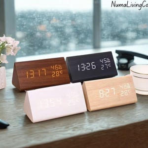 May include: Four wooden digital alarm clocks in various colors: dark brown, black, white, and light wood. Each clock displays the time, humidity, and temperature. The clocks are triangular in shape.