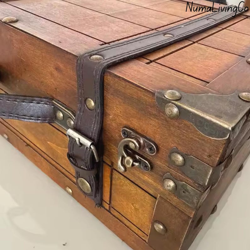 K&ouml;nnte beinhalten: Eine Holzkiste mit einem dunkelbraunen Lederriemen und Metallbeschl&auml;gen. Die Truhe hat eine rechteckige Form mit einem Klappdeckel und einem Metallverschluss. Das Holz hat eine warme, nat&uuml;rliche Oberfl&auml;che. Der Markenname "NumaLivingCo" ist sichtbar.