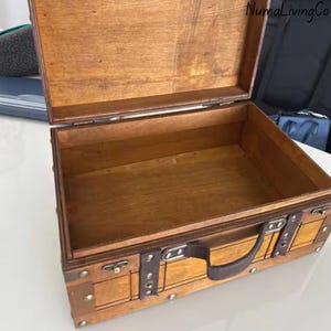 K&ouml;nnte beinhalten: Ge&ouml;ffnete Holzkiste mit braunem Ledergriff und Metallakzenten. Die Kiste ist rechteckig mit einem Klappdeckel und aus hellbraunem Holz gefertigt. Der Innenraum ist leer, und die Kiste scheint Vintage oder antik zu sein.