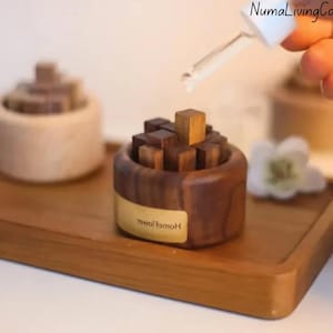 May include: A dark brown wooden essential oil diffuser, filled with small wooden blocks. A dropper dispenses liquid into the diffuser. The diffuser sits on a wooden tray, alongside a similar diffuser and a white flower.