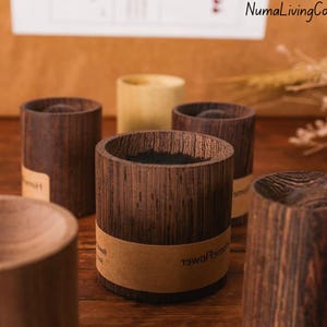 May include: Several cylindrical wooden containers with a dark brown finish. Each container has a natural wood grain pattern and a light brown label wrapped around the middle. The containers are of varying heights and are arranged on a wooden surface.
