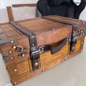 K&ouml;nnte beinhalten: Eine Holzkiste mit Vintage-&Auml;sthetik. Die Truhe ist braun mit dunkelbraunen Lederriemen und Griff. Sie verf&uuml;gt &uuml;ber Metalleckensch&uuml;tzer und Verschl&uuml;sse, die einen klassischen, antiken Stil hervorrufen. Die Truhe ist f&uuml;r die Aufbewahrung konzipiert.