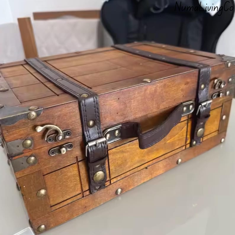 K&ouml;nnte beinhalten: Eine Holzkiste mit Vintage-&Auml;sthetik. Die Truhe ist braun mit dunkelbraunen Lederriemen und Griff. Sie verf&uuml;gt &uuml;ber Metalleckensch&uuml;tzer und Verschl&uuml;sse, die einen klassischen, antiken Stil hervorrufen. Die Truhe ist f&uuml;r die Aufbewahrung konzipiert.