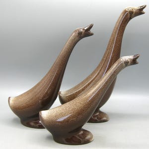 Könnte beinhalten: Ein Set aus drei Gänsefiguren aus Keramik mit einer glänzenden, braun gesprenkelten Glasur. Jeder Vogel ist mit einem langen Hals und geöffnetem Schnabel dargestellt und steht auf einem abgerundeten Sockel. Die Figuren variieren in der Höhe.