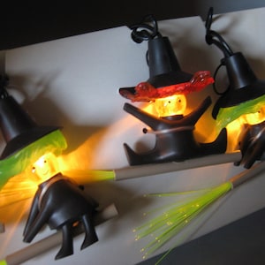 May include: A string of Halloween lights featuring witch figures riding broomsticks. Each witch has a black hat, a smiling face, and a black outfit. The lights are illuminated with a warm, yellow glow, creating a festive and spooky ambiance.