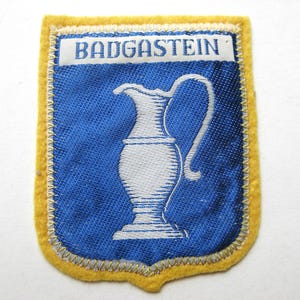 May include: A vintage patch with a blue background and a white pitcher design. The patch has a yellow border and the word "BADGASTEIN" in white letters across the top. The patch is likely a souvenir or emblem.