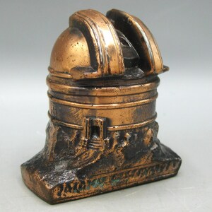 May include: A bronze-toned miniature observatory model. It has a domed roof with a slit, a cylindrical body, and a textured base. The design suggests a vintage or antique style, with intricate detailing throughout the structure.
