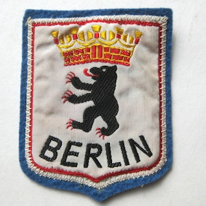 Könnte beinhalten: Gestickter Aufnäher mit dem Berliner Wappen. Das Design zeigt einen schwarzen Bären mit roten Krallen und Zunge, eine goldene und rote Krone und das Wort "BERLIN" in Schwarz. Der Aufnäher hat einen weißen Hintergrund, rote und weiße Nähte und einen blauen Rand.