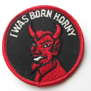 Pode incluir: Remendo bordado circular com um diabo vermelho sobre um fundo preto. O diabo tem chifres e uma expressão ameaçadora. O texto branco ao redor da borda diz "I WAS BORN HORNY". O remendo é bordado com costuras vermelhas.
