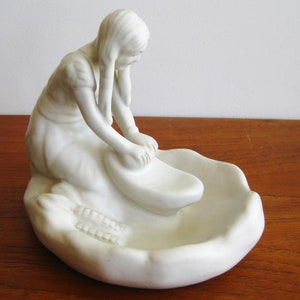 Könnte beinhalten: Eine mattweiße Porzellanfigur, die eine kniende Person beim Mahlen von Getreide auf einem traditionellen Stein-Metate darstellt. Die Skulptur hat eine glatte, unglasierte Oberfläche mit detaillierten Texturen an der Kleidung und dem Mahlwerkzeug und fängt einen Moment manueller Arbeit in einer monochromen, skulpturalen Form ein.