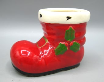 May include: A ceramic Christmas stocking-shaped container. The red boot has a white cuff with black accents and green holly leaves. The base is black, and gold accents are visible.