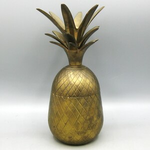 Peut inclure: Un récipient en laiton doré en forme d'ananas avec un couvercle amovible. Le corps présente une surface texturée à motif en losange, tandis que le sommet se compose de plusieurs feuilles pointues et dressées formant la couronne de l'ananas.