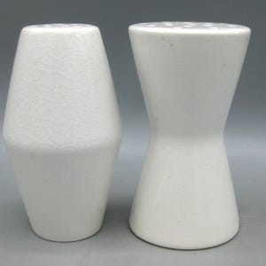 May include: A white ceramic mug with a black handle and the text "But First, Coffee" in a bold, black font. The mug is set against a blurred background. The mug appears to be approximately 10 cm tall.