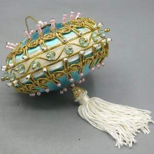 Peut inclure: Un ornement décoratif de forme ovale avec une base en satin bleu vif. La surface est ornée de tresses métalliques dorées, de bandes de ruban blanc, de paillettes bleu clair et de petites perles roses et blanches. Un pompon blanc est suspendu.