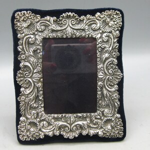 May include: A rectangular picture frame featuring an ornate, embossed silver-toned metal border with intricate scrollwork and floral motifs. The frame is set against a dark navy blue velvet backing, creating a high-contrast display for a photograph.