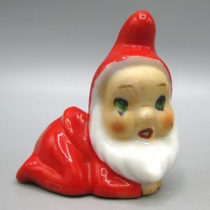Pode incluir: Uma figura de cerâmica de um personagem semelhante ao Pai Natal, com chapéu e fato vermelhos, barba branca e bochechas rosadas. A figura está em posição de engatinhar, com olhos verdes e uma expressão neutra. Uma decoração de Natal vintage.