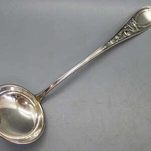 May include: A silver-toned metal ladle featuring a deep, circular bowl and a long, slender handle. The handle is decorated with an intricate, embossed scrollwork pattern and a smooth, oval-shaped panel at the very end of the stem.