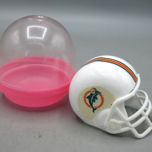 Op de afbeelding: Een witte miniatuur American footballhelm met oranje en witte strepen en een teamlogo. De helm staat naast een doorzichtige plastic capsule met een roze basis. De helm heeft een wit gezichtsmasker.