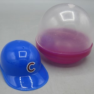 May include: A blue baseball cap with a white "C" logo next to a clear and pink plastic capsule. The cap has a curved brim and a rounded crown. The capsule is transparent with a pink base.