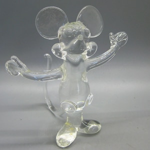 May include: A clear glass mouse figurine with outstretched arms and large ears. The mouse has a long tail and is standing on two feet. The figurine is made of transparent glass and is likely a decorative item.