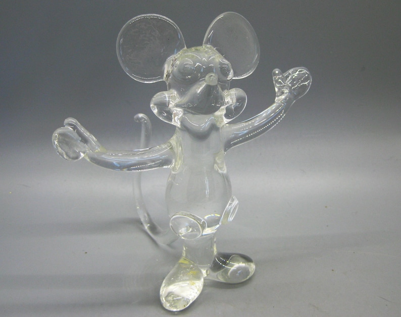 May include: A clear glass mouse figurine with outstretched arms and large ears. The mouse has a long tail and is standing on two feet. The figurine is made of transparent glass and is likely a decorative item.