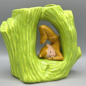 Pode incluir: Um vaso de cerâmica vintage em forma de tronco de árvore, pintado em verde brilhante com manchas escuras. Uma figura de esquilo marrom está aninhada dentro da abertura do tronco da árvore. O esquilo tem um rosto rosa claro e está em repouso.