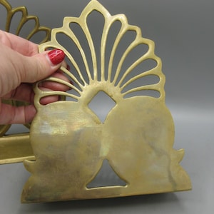 May include: A pair of vintage brass bookends. Each bookend has a fan-shaped top with a central diamond cutout and two rounded bottom sections. The brass has a warm, aged patina. The bookends are designed to stand upright.