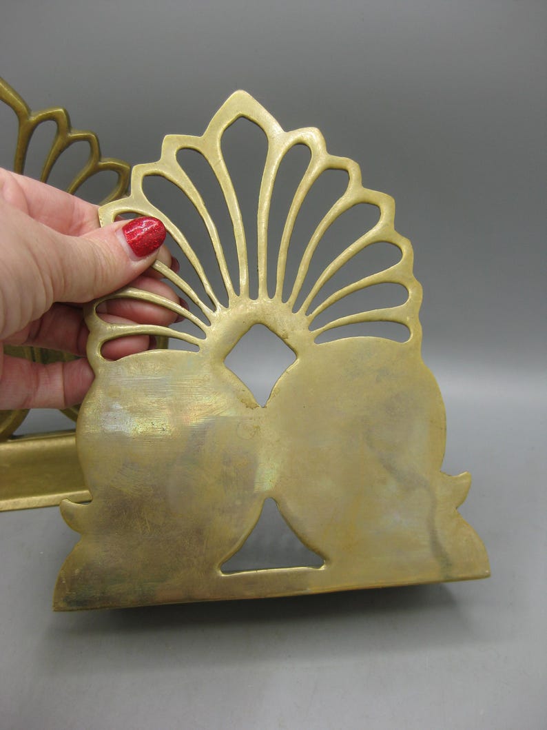 May include: A pair of vintage brass bookends. Each bookend has a fan-shaped top with a central diamond cutout and two rounded bottom sections. The brass has a warm, aged patina. The bookends are designed to stand upright.
