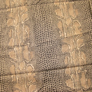 Könnte beinhalten: Ein Stoff mit Schlangenmuster. Das Design zeigt ein sich wiederholendes Muster aus dunkelbraunen und beigen Schuppen, wodurch ein strukturiertes Aussehen entsteht. Die Gesamtästhetik des Stoffes erinnert an Reptilienhaut.