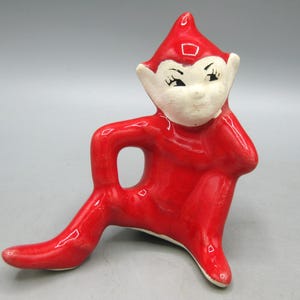 Pode incluir: Estatueta de cerâmica de um elfo sentado com corpo e chapéu vermelhos e rosto branco. O elfo tem olhos pretos e posa com um braço dobrado e a outra perna estendida. Um item decorativo vintage.