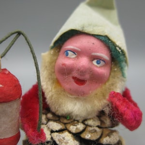 May include: A vintage Christmas ornament depicting a gnome holding a red lantern. The gnome has a pink face, blue eyes, and a white and green hat. The lantern is red with a blank label. The gnome's body is made of pinecones.