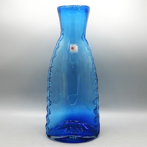 May include: A tall, blue glass vase with a textured, wavy design along the edges. The vase has a wide base that tapers to a narrower neck. A small label is visible near the top. The vase is approximately 12 inches tall.