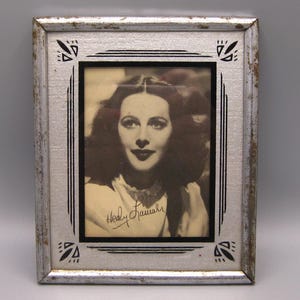 May include: A framed black and white photograph of a woman with dark hair and lipstick. The silver frame has decorative black accents and the woman's signature is visible. The photograph is vintage.