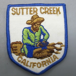 May include: Embroidered patch with a white background, blue border, and gold lettering reading "Sutter Creek California". The patch depicts a figure in a hat and blue and green clothing. The patch is likely a souvenir.