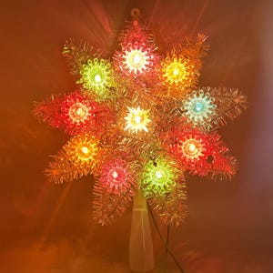 May include: A star-shaped Christmas tree topper made of metallic gold tinsel with multiple points. The ornament features several integrated, glowing light bulbs in various colours including red, yellow, green, and blue, with a clear plastic base.