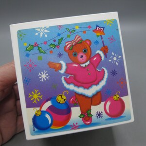 May include: A square, white box with a colorful Christmas-themed illustration. The image features a dancing teddy bear in a pink dress, holding a star wand, with Christmas ornaments and snowflakes. The background transitions from blue to purple.